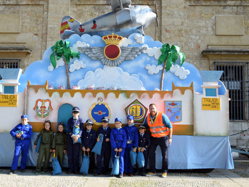 Una imagen de una carpa festiva con un diseño de avión y corona, junto a niños vestidos como soldados y adultos en uniforme. La carpa está decorada con banderas y un cartel que dice "Prisión". En el fondo, se puede ver una pared de piedra con ventanas.