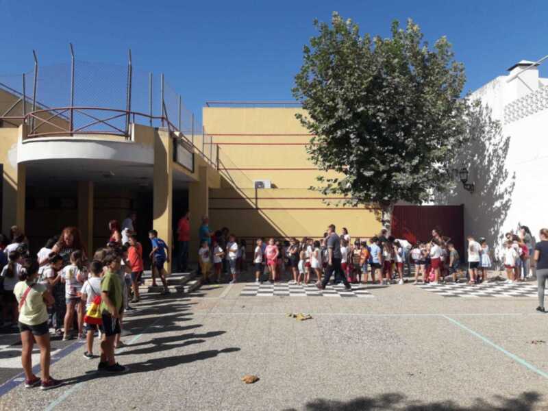 Un grupo de niños y adultos están esperando en fila frente a una escuela con un edificio amarillo y un árbol grande. El cielo es azul claro, indicando buen tiempo.