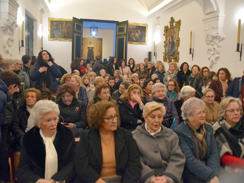 Una multitud de personas sentadas en filas, mirando hacia adelante. La imagen muestra una sala con paredes blancas y decoración clásica, incluyendo cuadros y molduras. La gente está vestida formalmente, lo que sugiere una reunión o evento importante.