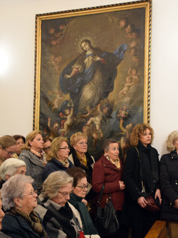 Una multitud de personas se reúne en un salón con una pintura religiosa de la Virgen María y ángeles en el fondo. La escena parece ser un evento formal o religioso, con las personas vestidas de manera elegante.