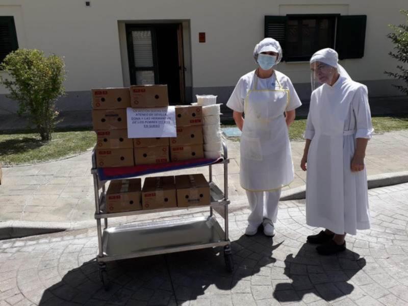 Dos personas vestidas con uniformes blancos y máscaras protectoras sostienen una carretilla de cajas. La escena se encuentra frente a un edificio con ventanas y una puerta, en un entorno urbano con césped y vegetación.
