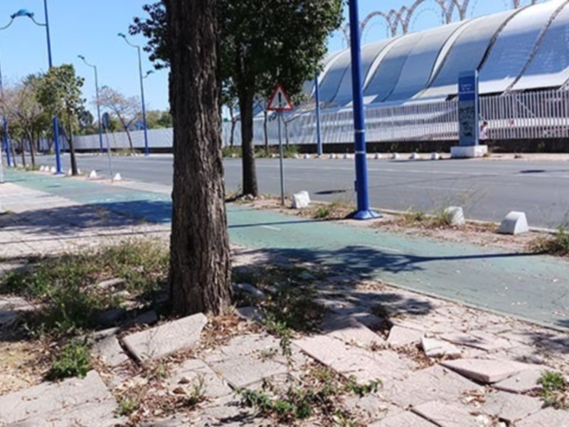 Área urbana con árboles, sendero peatonal y edificio de techos curvos en el fondo.