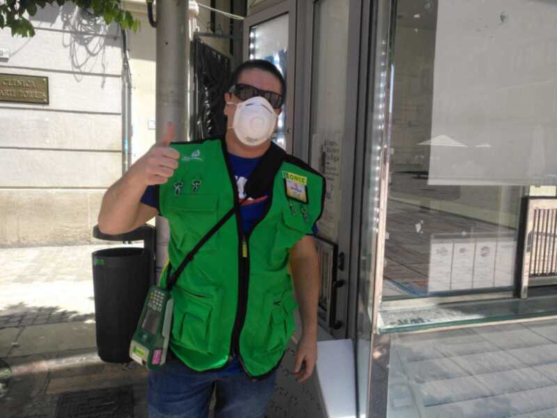 Un hombre con una chaqueta verde y un chaleco de trabajo, posando con la mano levantada. Detrás de él se puede ver una ventana de cristal y un edificio con un letrero.