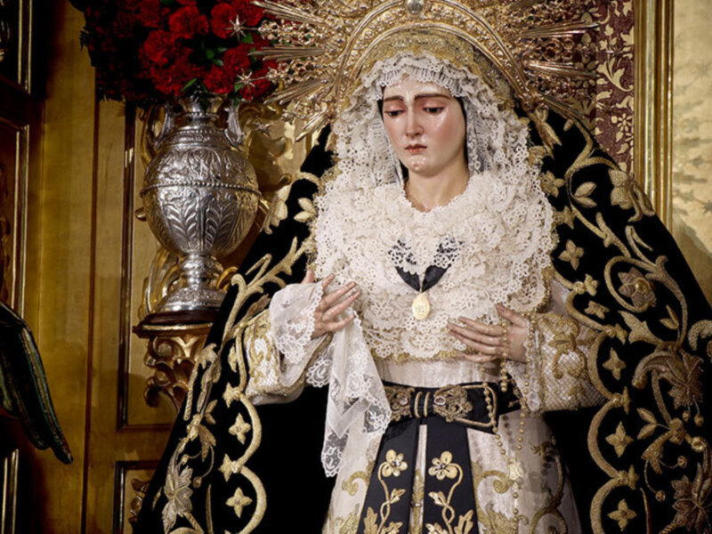 Imagen de una Virgen María vestida con trajes tradicionales, incluyendo un manto negro y dorado. La imagen está en una iglesia con detalles arquitectónicos y flores rojas al fondo.