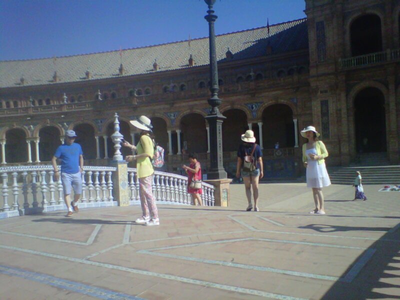 En la Plaza de España, Sevilla, se observan varios visitantes disfrutando del entorno. La arquitectura es impresionante con sus grandes arcadas y detalles en azulejo. La gente está vestida informalmente, algunas con sombreros de ala ancha para protegerse del sol. La imagen captura el ambiente tranquilo y agradable de un día soleado en esta famosa plaza.