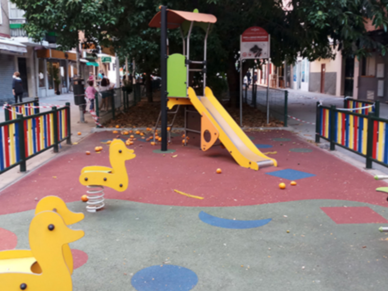 Un parque infantil con juegos de madera y una gran escalera. La zona está rodeada por un camino peatonal con bancos de madera y una cerca de metal. En el fondo, se pueden ver árboles y edificios.