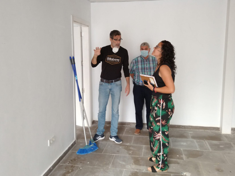 En una habitación vacía, tres personas discuten sobre un proyecto. La persona central, vestida con una camiseta negra y pantalones azules, sostiene un libro abierto. A su izquierda, una persona con camisa verde y pantalones de cuadros sostiene un bolígrafo. A la derecha, una persona con pantalones estampados y sandalias sostiene un portátil. La pared es blanca, con una puerta abierta a la izquierda y un piso de baldosas grises.
