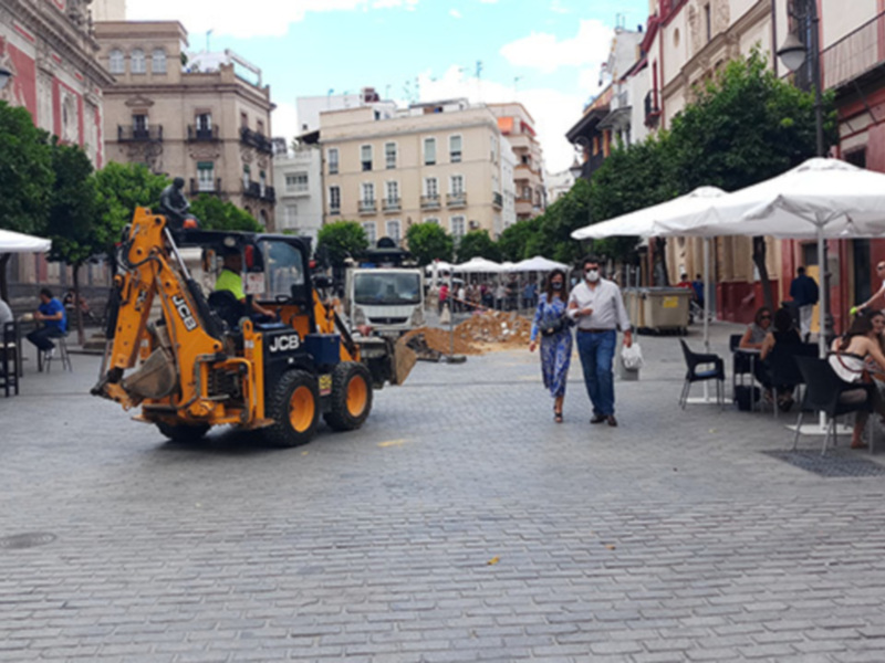 Un excavador amarillo se mueve por una calle adoquinada, mientras personas caminan y otros trabajan en el fondo. La imagen muestra una mezcla de actividad urbana con elementos de construcción y vida cotidiana en un entorno arquitectónico.
