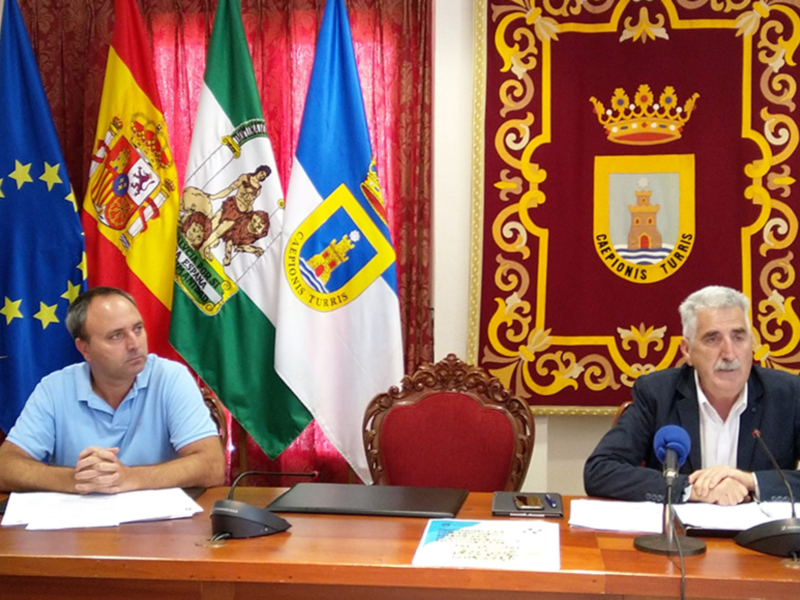Dos hombres sentados en una mesa con banderas españolas y europeas, un documento sobre la mesa y micros para comunicaciones.