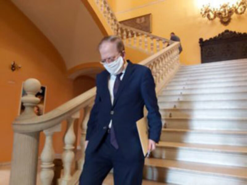 Un hombre en traje azul y máscara blanca camina por una escalera de mármol blanco en un entorno amarillo. La decoración clásica y el ambiente formal sugieren una escena de oficina o evento importante.