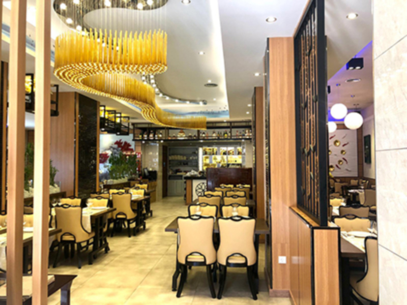 Interior moderno de un restaurante con mesas redondas y sillas tapizadas en beige, iluminado por una lámpara de araña dorada y decorado con plantas.
