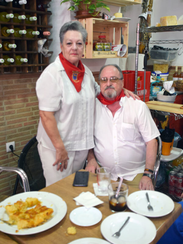 Un hombre y una mujer sonríen en una cocina, posando junto a una mesa con platos de comida y bebidas.