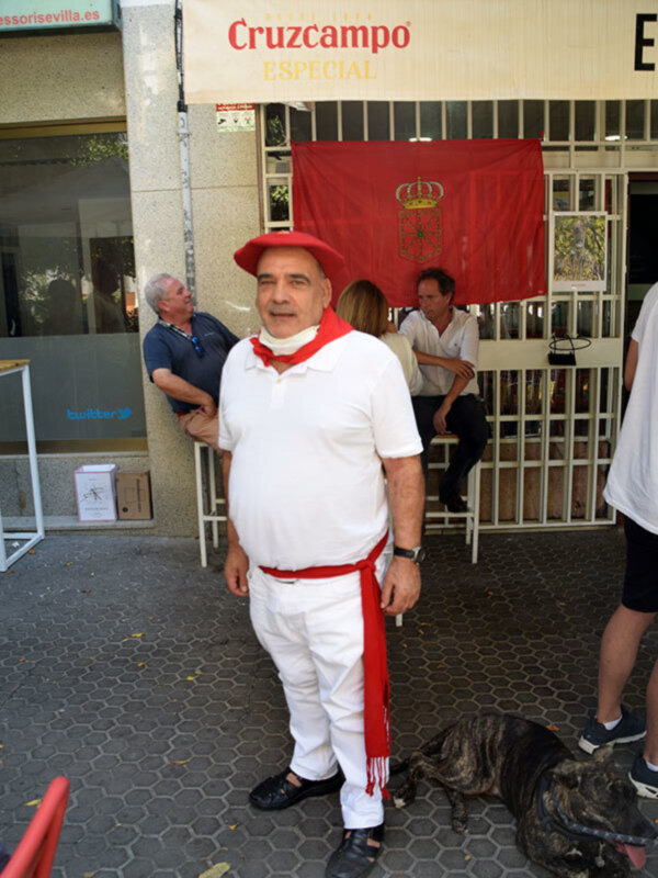 Un hombre vestido con traje tradicional español, incluyendo sombrero rojo y bufanda roja, posa frente a un edificio con una bandera roja. Detrás de él, se observa una persona sentada y otros dos individuos en la calle. En el fondo, hay un anuncio de Cruzcampo Especial y una ventana con persianas.