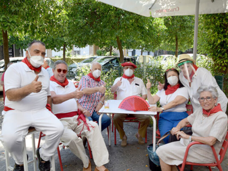 Un grupo de personas mayores se sientan en sillas rojas bajo un paraguas blanco. Algunos llevan gorros rojos y bufandas, mientras que una mujer con gafas de sol muestra un gesto de apoyo. La escena parece ser una reunión o evento en el aire libre, con árboles verdes al fondo.