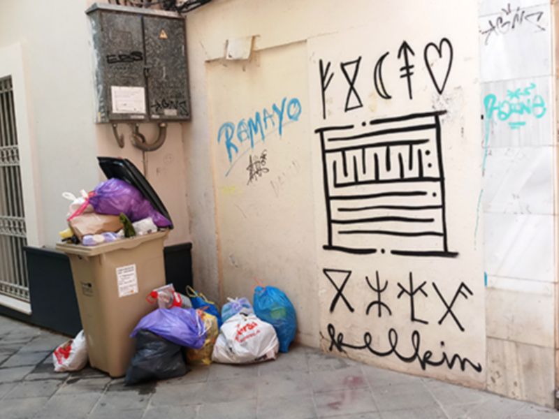 Se observa una pared con grafitis y un contenedor de basura lleno. El grafiti incluye letras y símbolos que parecen ser parte de un diseño artístico. La pared también tiene pegatinas y etiquetas colgadas en diferentes lugares.