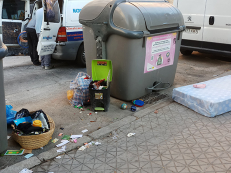 Un contenedor de basura lleno de residuos, incluyendo un pañuelo de papel y una caja de cartón, junto a otros desechos en el suelo. Un cartel informativo está pegado al contenedor, y una camioneta blanca con el logo de Peugeot está estacionada en el fondo.