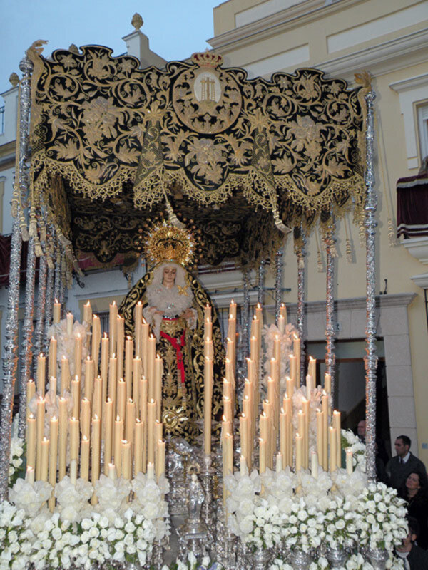 Imagen de una procesión religiosa con una imagen venerada rodeada por velas encendidas y flores blancas. El entorno muestra detalles arquitectónicos de un edificio histórico, con una estructura ornamental en el techo.