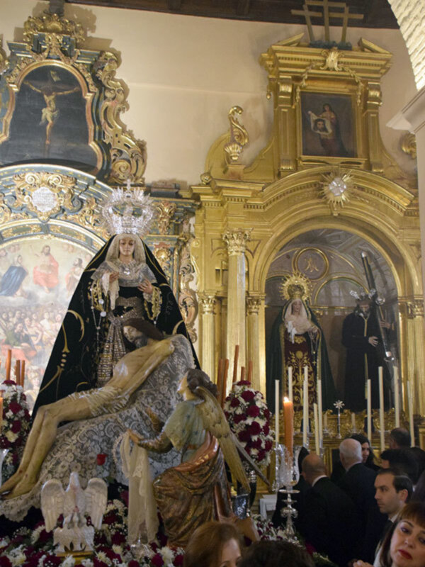 Una imagen de una iglesia con altares dorados, estatuas religiosas y flores. La imagen muestra una escena de celebración o procesión, con personas observando la estatua central. La decoración es elaborada y colorida, destacando el ambiente festivo de la iglesia.