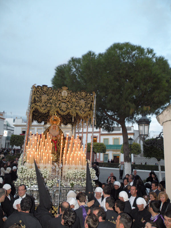 Una procesión religiosa con una imagen venerada en un carro adornado, rodeada de fieles vestidos con trajes tradicionales y sombreros negros. El cielo es claro, con árboles en el fondo y edificios blancos alrededor.