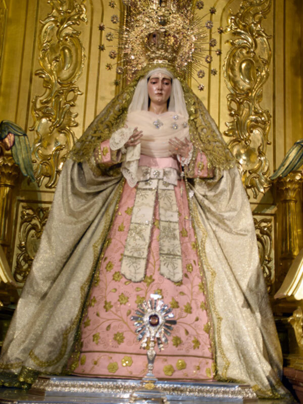 Imagen de la Virgen María vestida con un manto rosa y dorado, con una corona de diamantes y un escudo en la frente, en una iglesia con decoración dorada en el fondo.