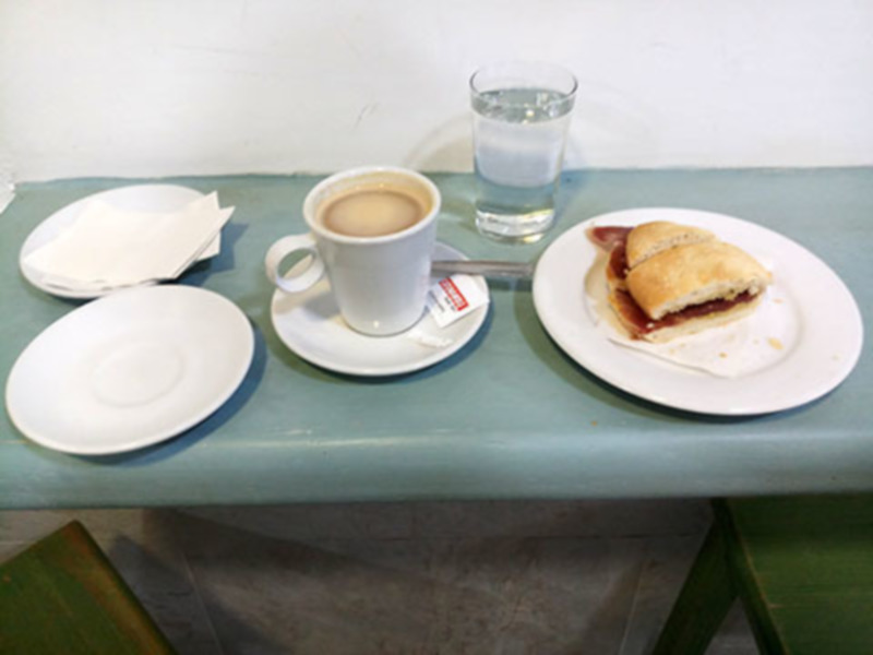 Se observa una mesa de color azul claro con dos platos, uno vacío y otro con un sándwich cortado en mitades. A la derecha del plato con el sándwich, hay una taza blanca de café y un vaso de agua. La mesa está sobre un suelo de baldosas grises, y al fondo se puede ver una pared blanca.