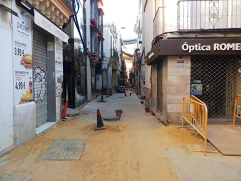 Una calle estrecha y desierta con varios locales comerciales. A la izquierda, se puede ver una tienda de comida rápida con carteles en español y francés. A la derecha, hay una tienda llamada "Optica Romeo" con un letrero en español. La calle está bordeada por edificios de dos pisos y hay algunas señales de tráfico. La imagen transmite un ambiente urbano típico, con tiendas y edificios típicos de una ciudad.