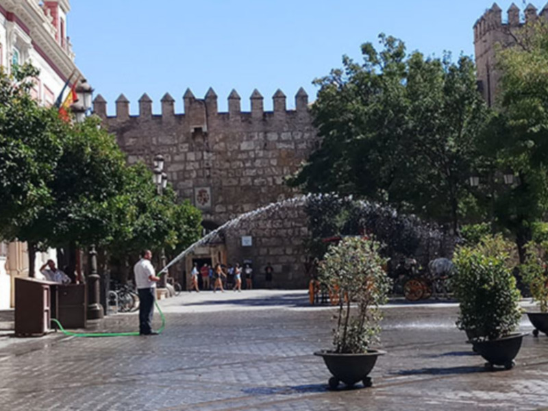 Plaza histórica con fuente de agua y murallas medievales en un día soleado.