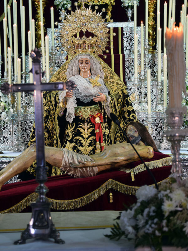 Imagen de una Virgen María en trono, rodeada por velas y flores. En el frente, un Cristo crucificado sobre una silla de terciopelo rojo. Detalles dorados y cristales en la decoración del trono.