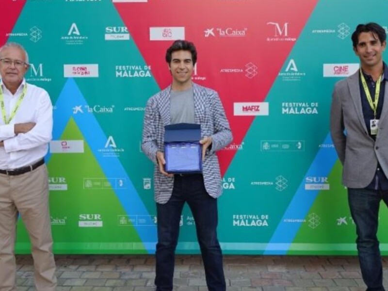 Festival de Málaga 2023. Un joven sostiene un premio mientras dos personas lo observan con orgullo. El fondo es verde y blanco con logos de la organización del festival.