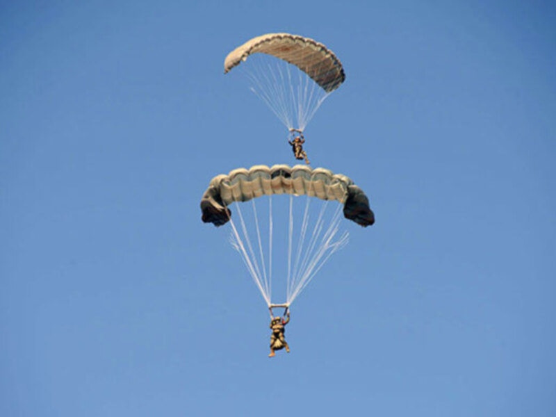 Un paracaidista en traje militar desciende con un paracaídas de color beige y negro. El cielo azul claro proporciona un fondo perfecto para este espectáculo aéreo.