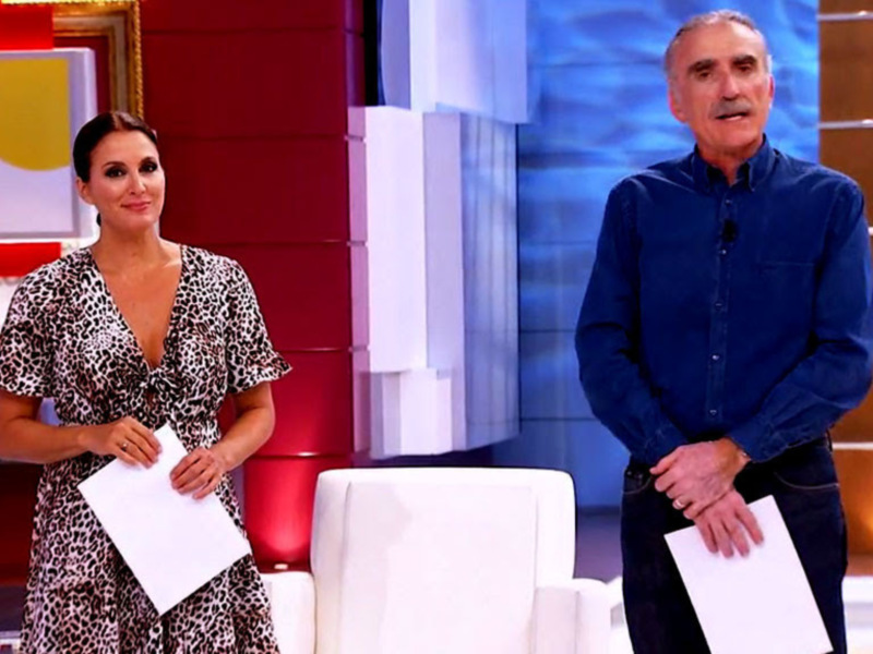 En el escenario de un programa televisivo, una presentadora con vestido estampado y cabello largo presenta a un hombre en camisa azul, ambos sujetan papeles. El fondo es rojo con elementos gráficos y una luz dorada en el suelo.