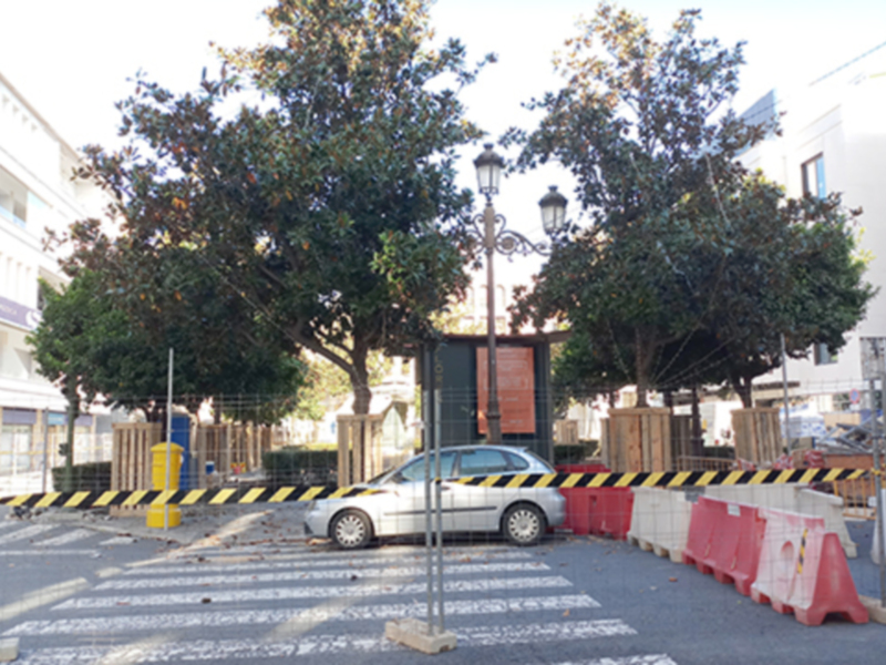 Un coche blanco está estacionado en una calle con un poste de luz y dos árboles grandes al fondo. Hay barreras amarillas y rojas en el camino, indicando un área de trabajo o construcción. El paisaje urbano incluye edificios blancos y una calle con líneas de peatones.