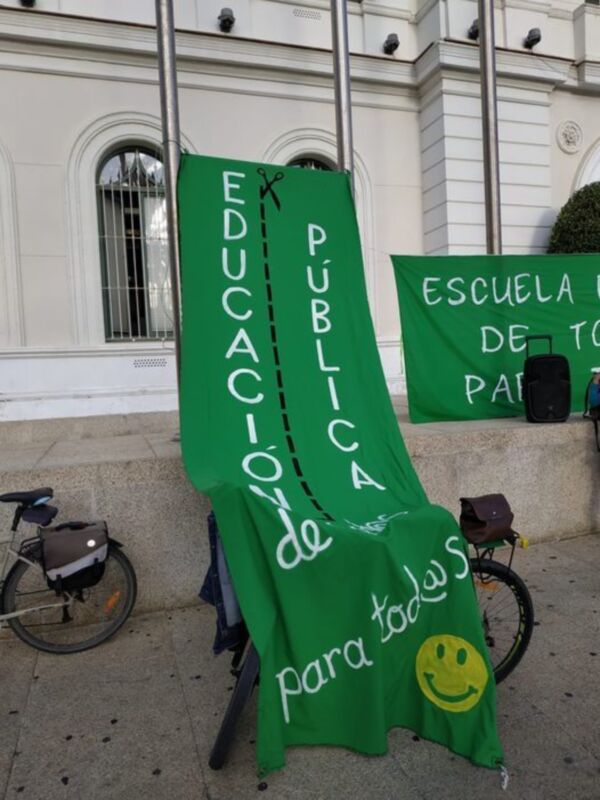 Pié de foto: "Educación pública para todos. Pancartas verdes con mensaje y smiley en frente de un edificio blanco."