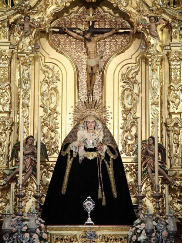 Imagen de la Virgen María en un altar dorado con detalles artísticos. La escena incluye una cruz y figuras de ángeles en el fondo.