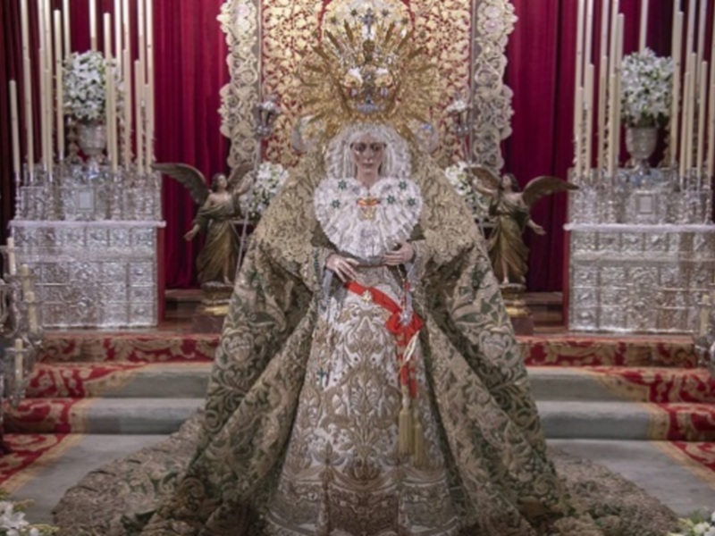 Una imagen de una figura vestida con un manto y corona en un altar, rodeada por flores y velas.