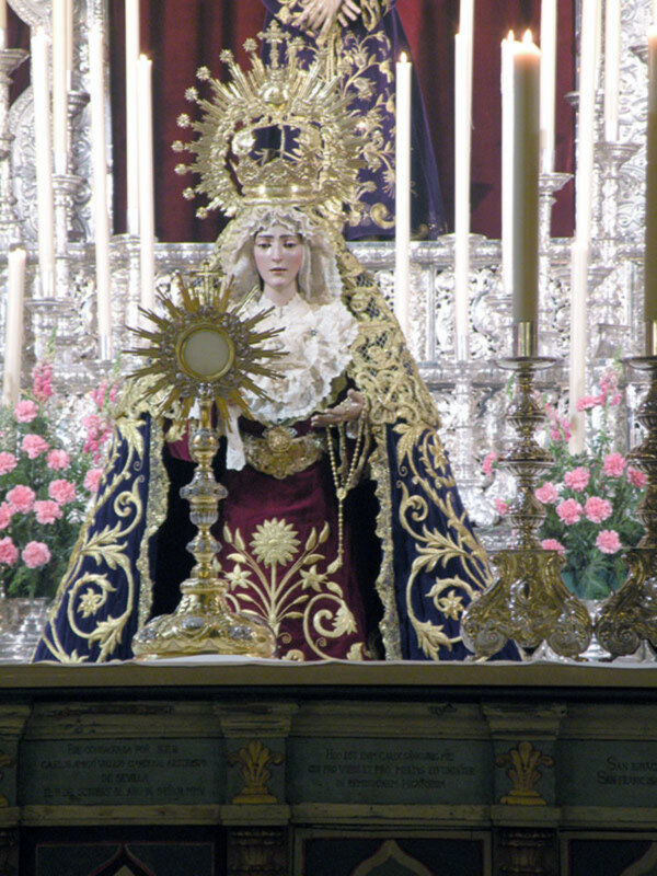 Imagen de una Virgen María vestida con un manto dorado y rojo, rodeada por flores rosas en un altar de iglesia. La imagen está iluminada con velas y hay inscripciones en el fondo.
