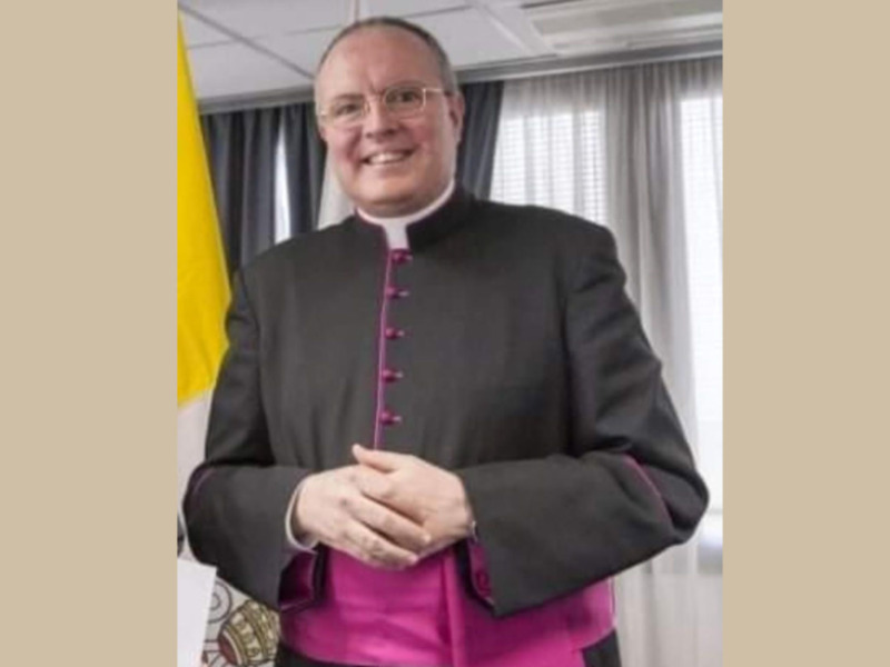 El sacerdote está posando con una sonrisa, vestido con un hábito negro y rosa. La imagen incluye una bandera en el fondo, lo que sugiere un contexto religioso o oficial.