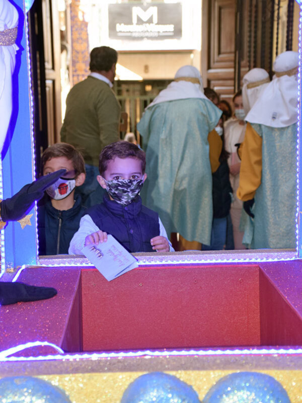 Un niño con una máscara de Halloween en un escaparate navideño. Detrás, figuras vestidas con trajes de reyes magos y un cartel con la letra "M". La imagen está iluminada con luces de colores.