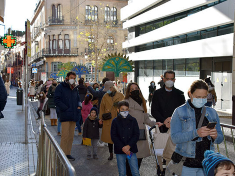 Una multitud de personas camina por una calle con edificios históricos a ambos lados. Algunas personas llevan máscaras faciales, mientras otros miran sus teléfonos. La escena parece ser de una ciudad vieja con arquitectura clásica y un cielo despejado.