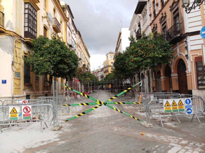 Calle bloqueada con barreras amarillas y negras, señalizadas con carteles de advertencia. Edificios históricos a ambos lados, árboles con flores en los balcones.