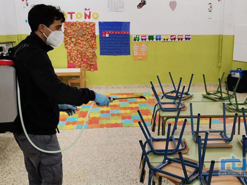 Un hombre desinfecta una escuela con un equipo de desinfección. La escuela tiene sillas y mesas, una alfombra con un diseño de rompecabezas y carteles en la pared. La imagen muestra el cuidado sanitario en un entorno educativo.