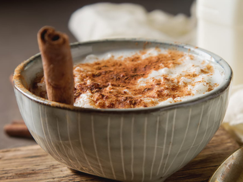 Un tazón de café con leche espeso, adornado con una canela y una cucharilla de azúcar blanca. La imagen se centra en el detalle de la textura del café y la espuma blanca, con un fondo suave y neutro.