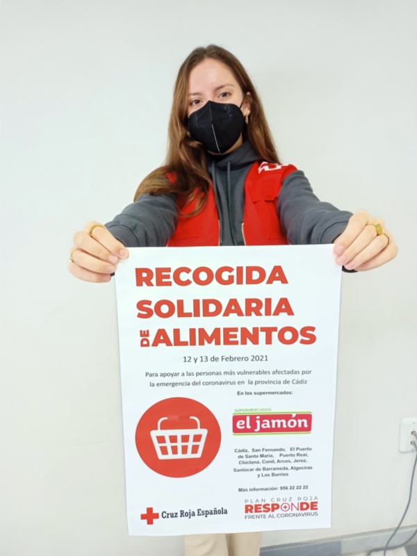 Una mujer con una máscara negra sostiene un cartel blanco que anuncia una recogida solidaria de alimentos para Cáritas en Febrero 2021. La imagen promueve la colaboración para ayudar a las personas más vulnerables afectadas por la emergencia del coronavirus.