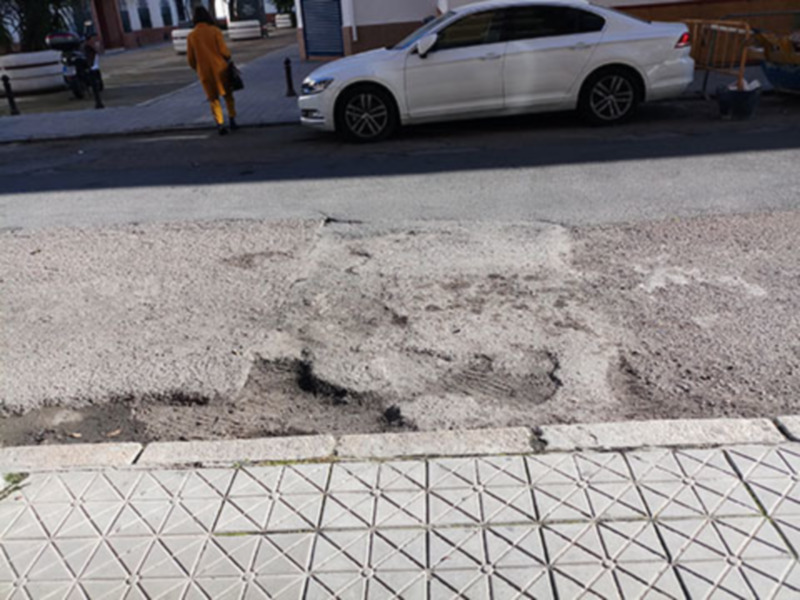 Un aparcamiento con una placa de estacionamiento marcada en el asfalto, que parece estar deteriorada o dañada. La placa está parcialmente oculta por la pintura de un vehículo blanco estacionado detrás. La calle es de asfalto claro y el entorno parece ser urbano, con una persona caminando en la calle de fondo.