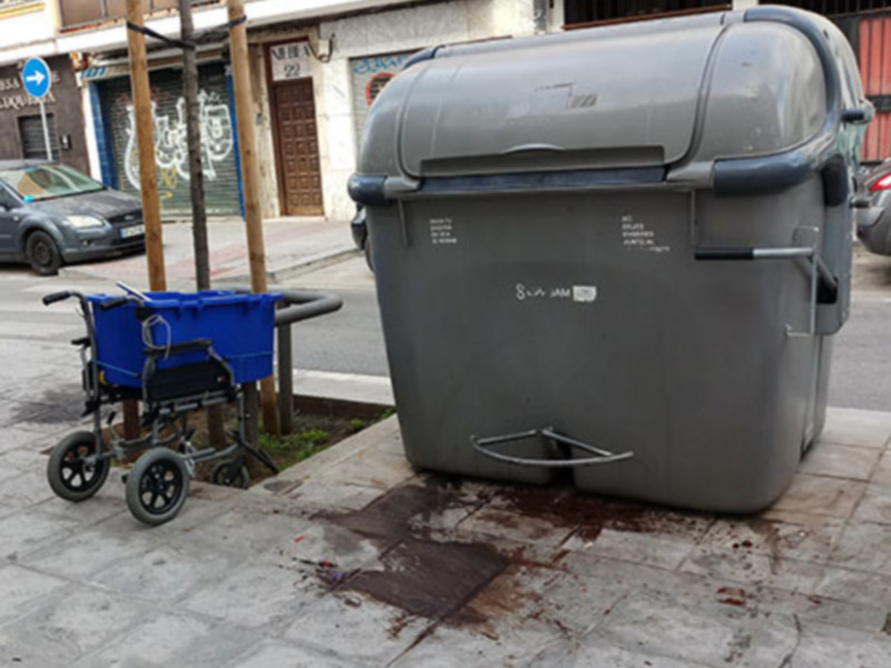 Un carrito de bebé azul y una basura gris en la calle, con un edificio de fondo y una señal de dirección azul con una flecha hacia la izquierda.