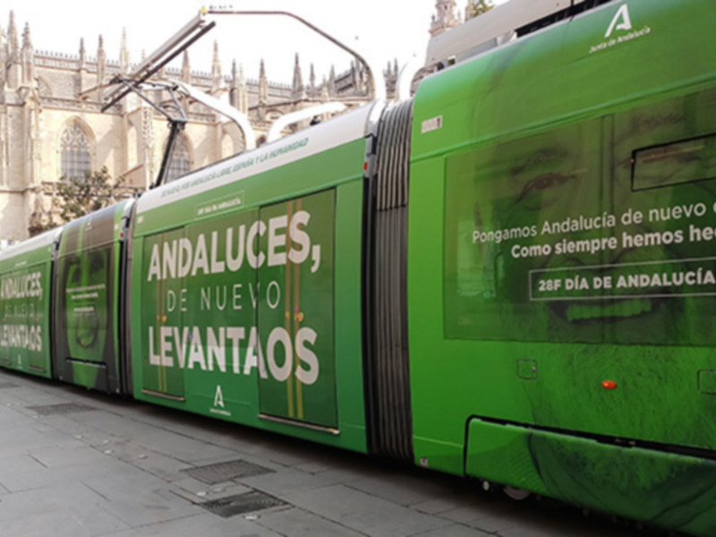 Tren verde con anuncios en español, frente a una catedral.
