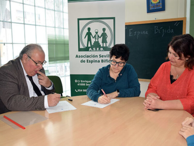 Se observa a tres personas sentadas en una mesa, dos de ellas firmando documentos mientras la tercera observa. La pared detrás de ellos tiene un cartel con el nombre "Asociación Sevilla de Espina Bífida" y una escuela. La mesa tiene un marcador rojo y un bloc de notas blanco.
