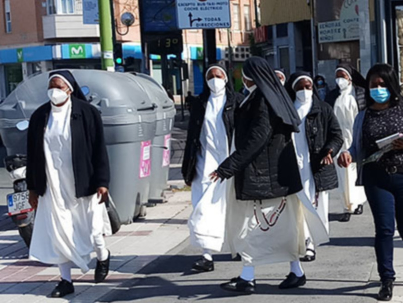 Una imagen de varias personas caminando por una calle, algunas con vestimentas religiosas y máscaras faciales. En el fondo se observan edificios, carteles de publicidad y un par de autos.