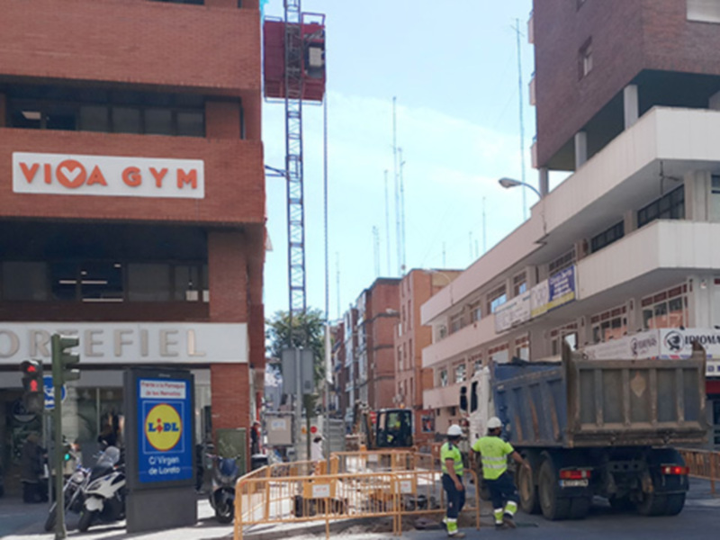 Se observa un entorno urbano con edificios de varios pisos, una construcción en proceso y una camioneta de trabajos. En el fondo hay un edificio con la señal "Viva Gym" y otro con una tienda de ropa. La escena parece estar en un área comercial o industrial, con trabajadores en cascos y una motocicleta estacionada.