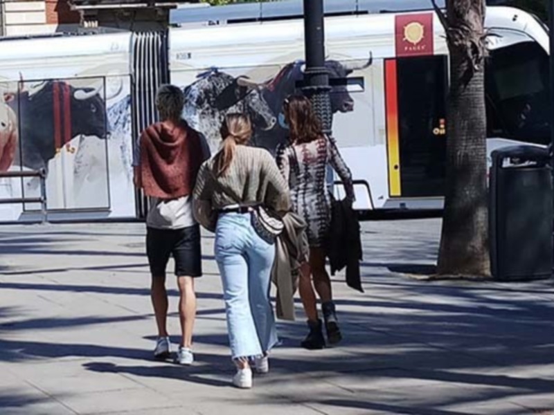 Tres personas caminan por una calle con un autobús en el fondo. La imagen muestra a dos mujeres y un hombre, todos vestidos de manera casual. El ambiente parece ser urbano con sombras caídas en el suelo, indicando que es un día soleado.